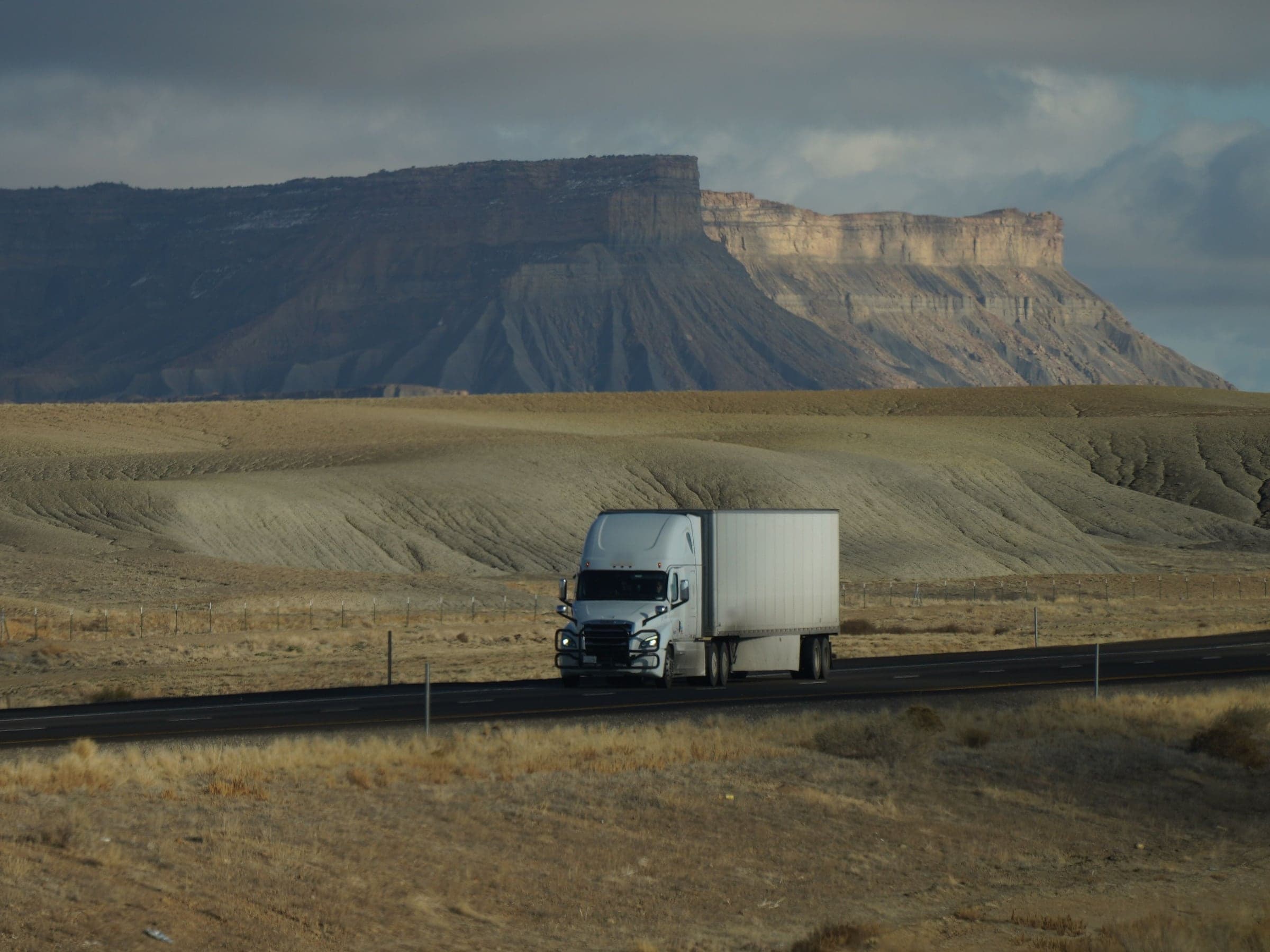 White semi-truck with an enclosed dry van trailer crossing a desert highway under a stormy sky